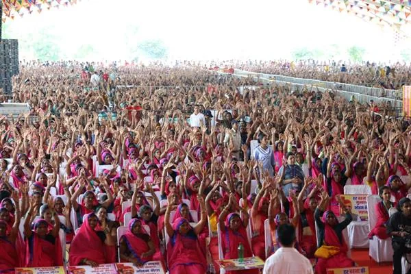 पंडाल में लाल और गुलाबी साड़ियों में बैठी बड़ी संख्या में महिलाएं हाथ उठाए हुए हैं