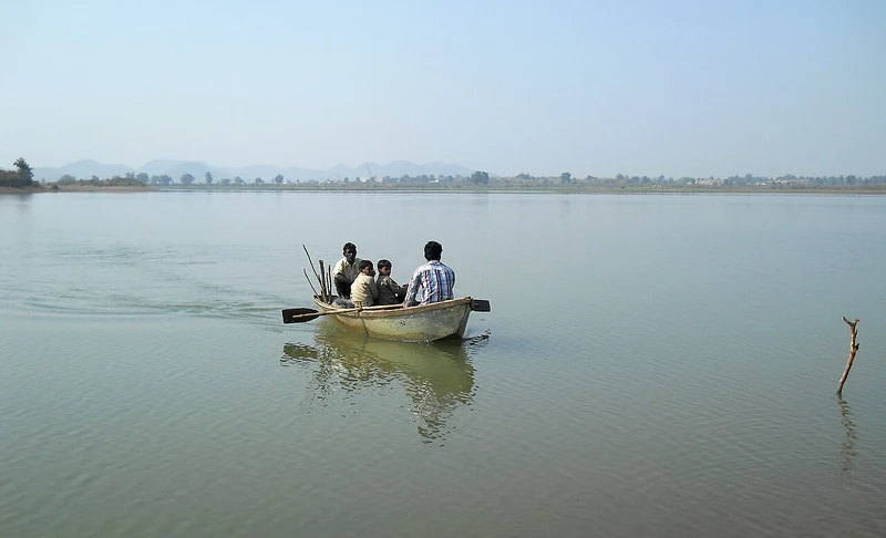 मध्य प्रदेश में पानी पर चलती छोटी लकड़ी की नाव में बैठे लोग, दूर किनारा और पहाड़ियाँ